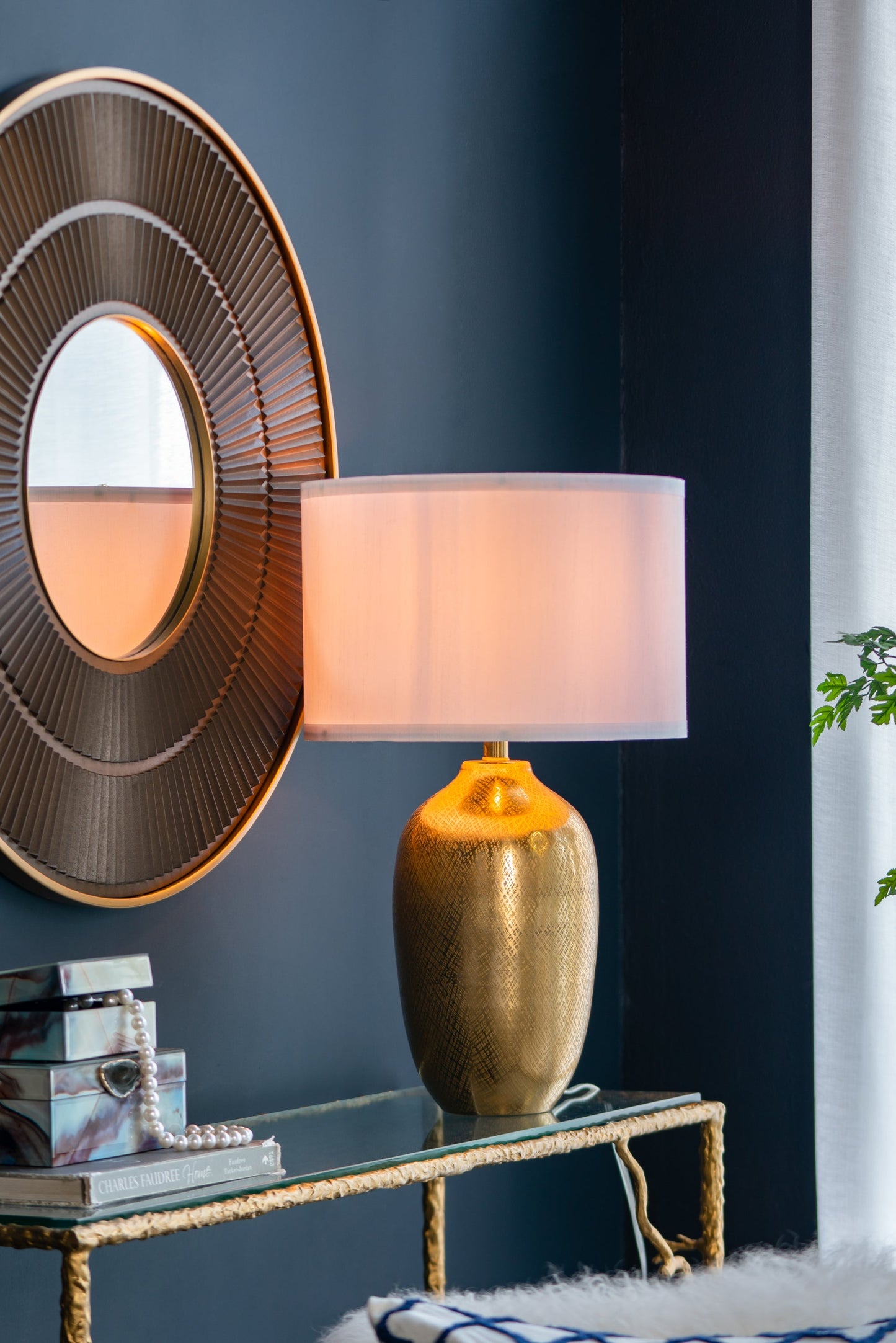 Decorative lamp with a gold base and pink shade on a glass table against a dark blue wall.