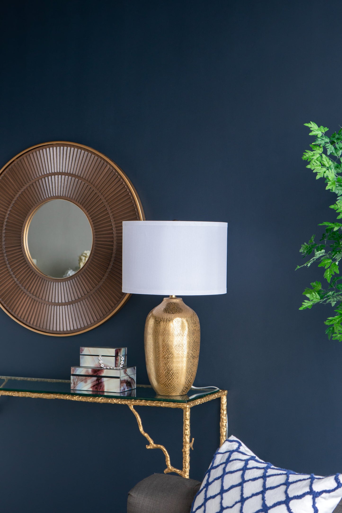 Gold lamp with white shade on a dark blue wall with decorative mirror and console table.