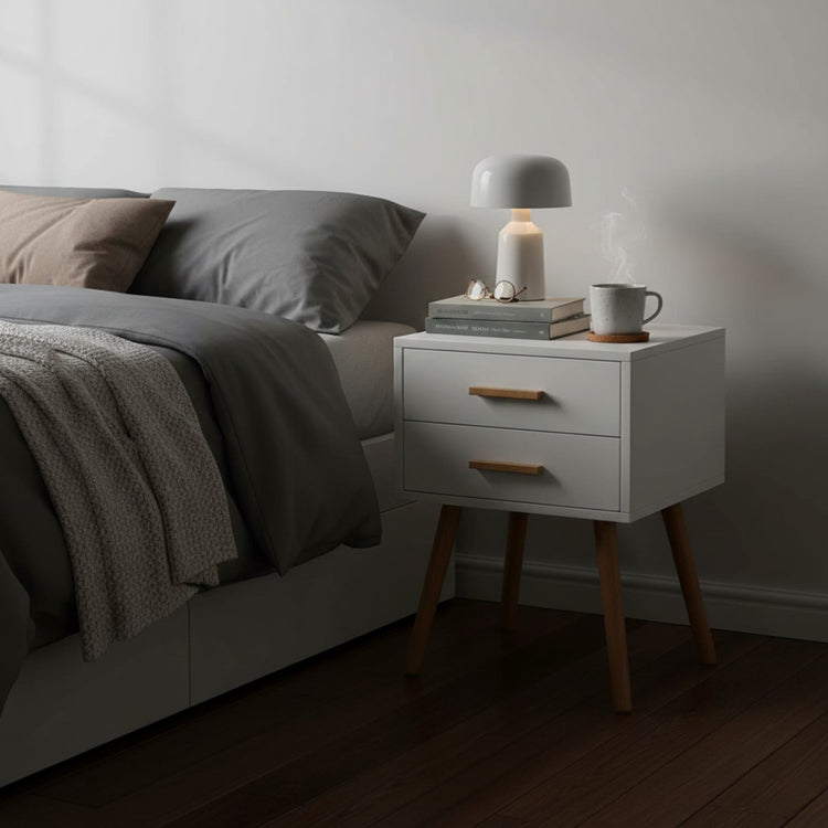 White piano-finish two-drawer end table with wooden legs beside a bed, topped with a lamp and books.