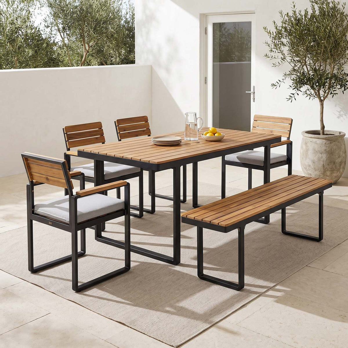 6-piece acacia outdoor dining set with bench on a sunlit limestone terrace, wide view