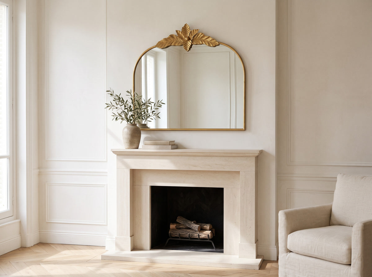 Gold arched mirror with leaf crest hung above a cream stone mantel beside a vase of olive branches