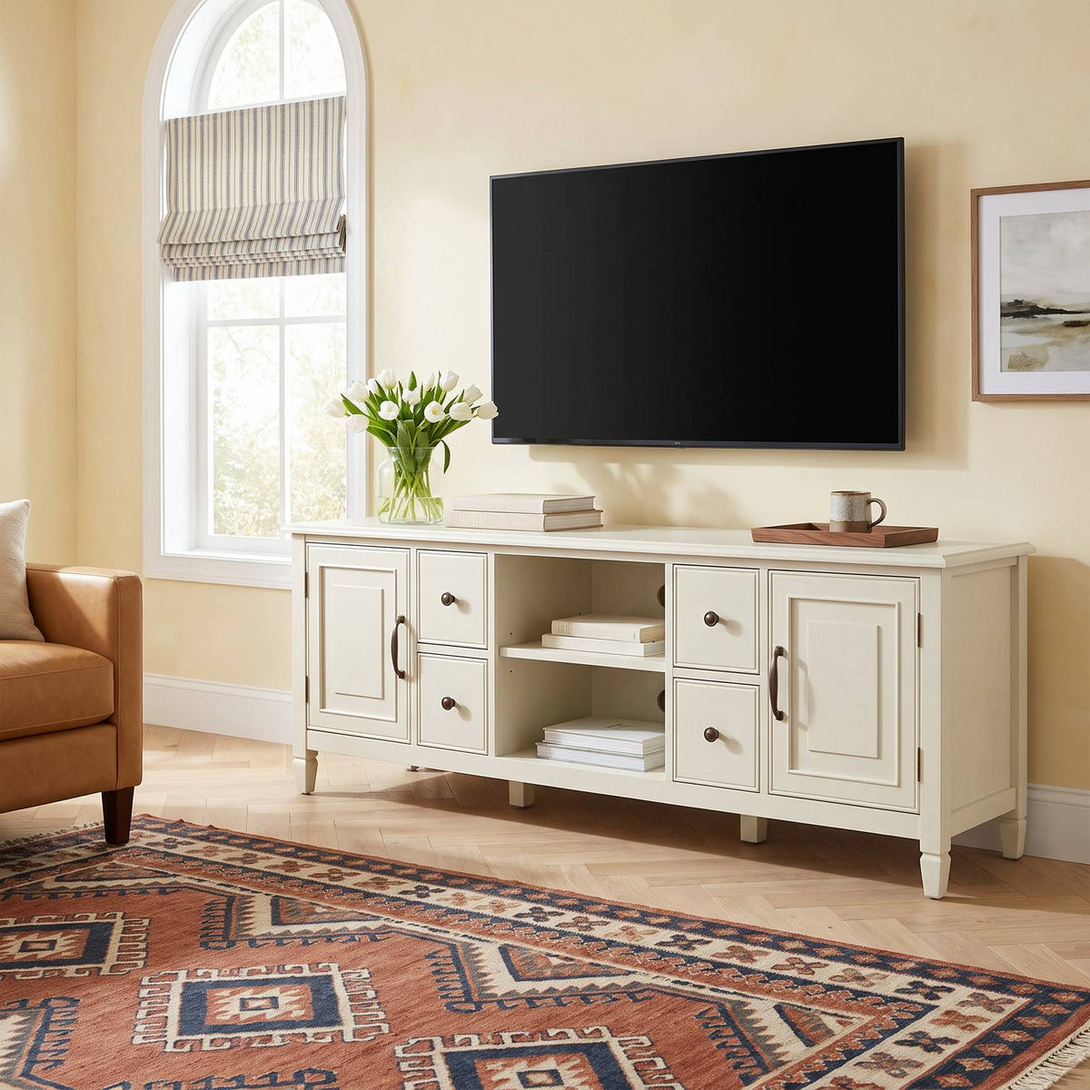 Antique white tv stand under a wall-mounted TV in a sunlit den with an arched window, tulips, books and a tan armchair
