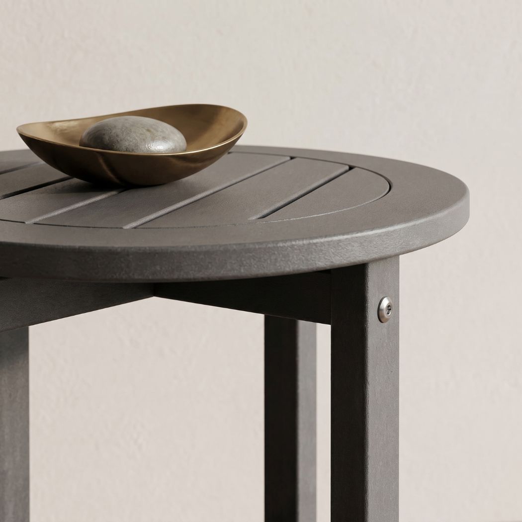 A close-up of a round gray slatted side table with a small metallic bowl and stone on top, set against a plain light beige wall.