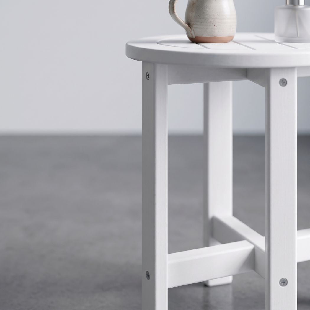 A close-up of a white wooden side table with a slatted top and open frame legs, shown in a minimalist room setting with a gray floor and a ceramic pitcher on the tabletop.