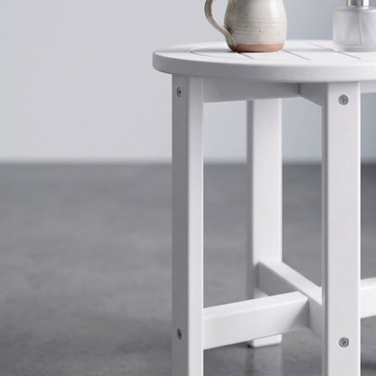 A close-up of a white wooden side table with a slatted top and open frame legs, shown in a minimalist room setting with a gray floor and a ceramic pitcher on the tabletop.