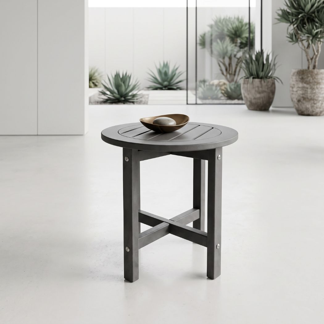 A round dark gray slatted side table with a small metallic bowl on top, set in a bright modern interior with potted plants in the background.