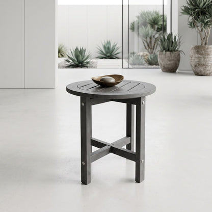 A round dark gray slatted side table with a small metallic bowl on top, set in a bright modern interior with potted plants in the background.