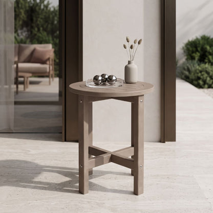 A round outdoor side table with a light taupe finish, styled with a small vase and metallic ornaments on a patio.