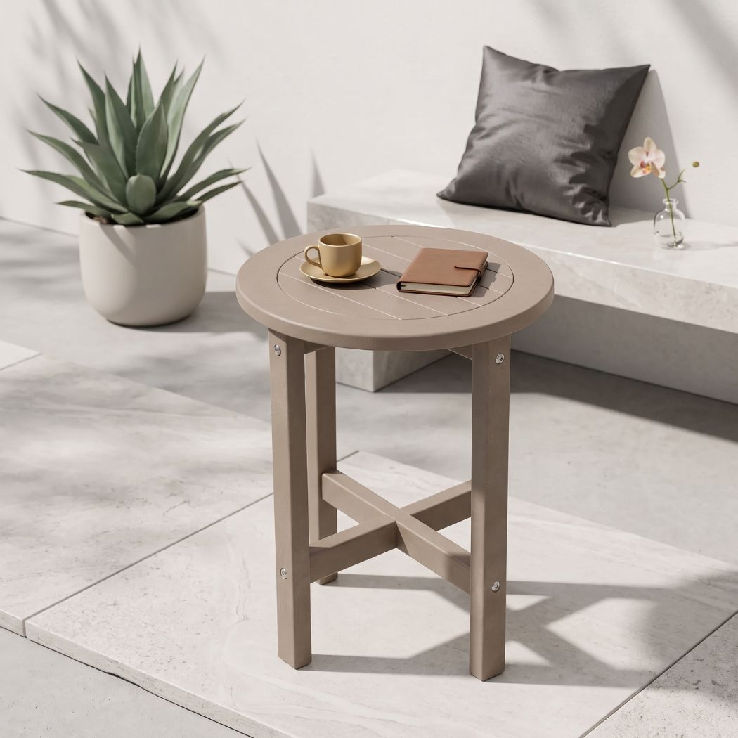 A round outdoor side table with a light taupe finish, styled in a modern patio setting with a coffee cup, notebook, potted plant, and bench in the background.