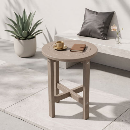 A round outdoor side table with a light taupe finish, styled in a modern patio setting with a coffee cup, notebook, potted plant, and bench in the background.