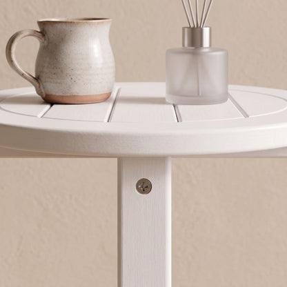 A white slatted round side table shown from above, with a speckled beige ceramic mug on the left and a frosted glass reed diffuser on the right, against a warm beige wall.