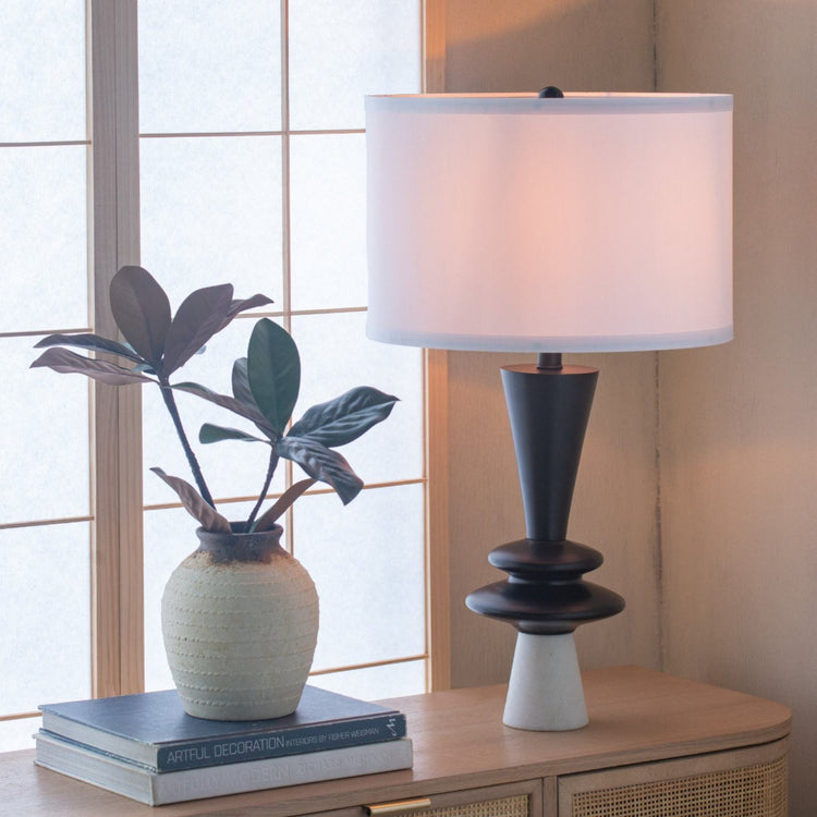 Aluminum Table lamp with white shade and black base on a wooden surface next to a plant and books.
