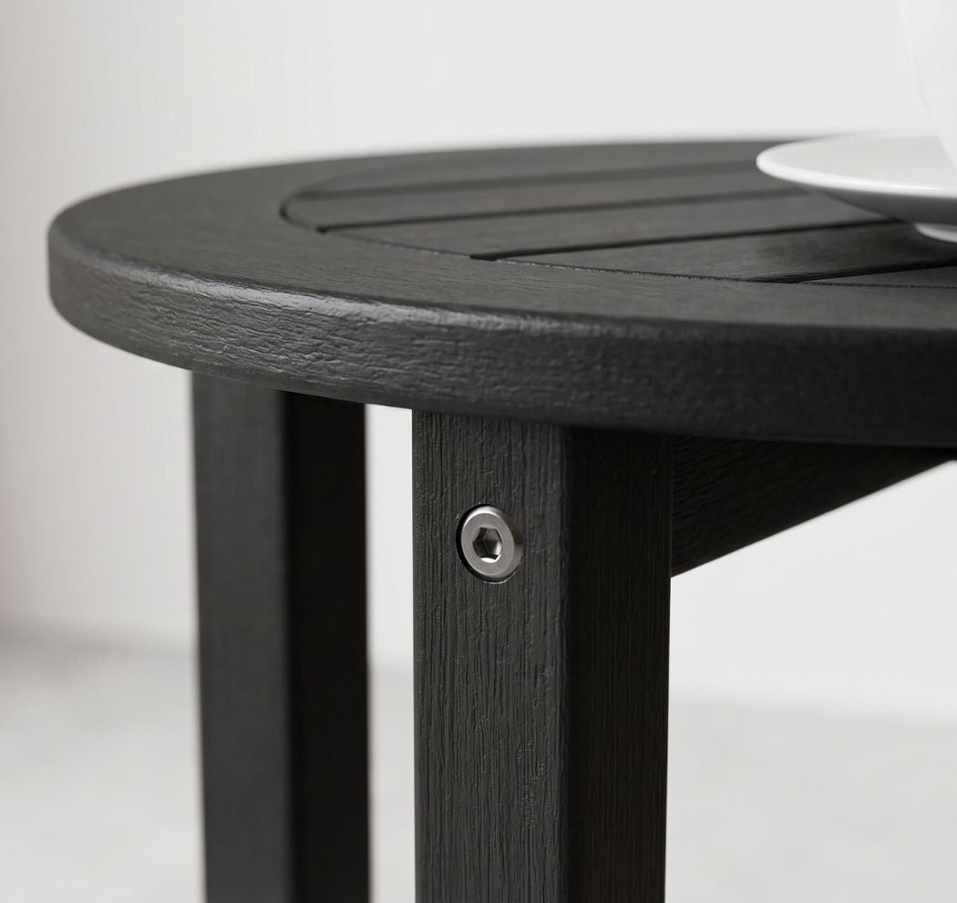 Close-up of a round black slatted table with a visible metal screw fastener on the leg, set against a light neutral background with a white plate partially visible in the top right.