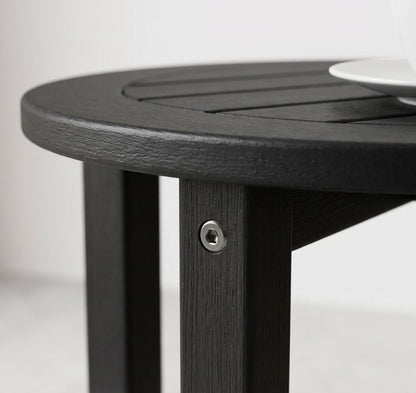 Close-up of a round black slatted table with a visible metal screw fastener on the leg, set against a light neutral background with a white plate partially visible in the top right.