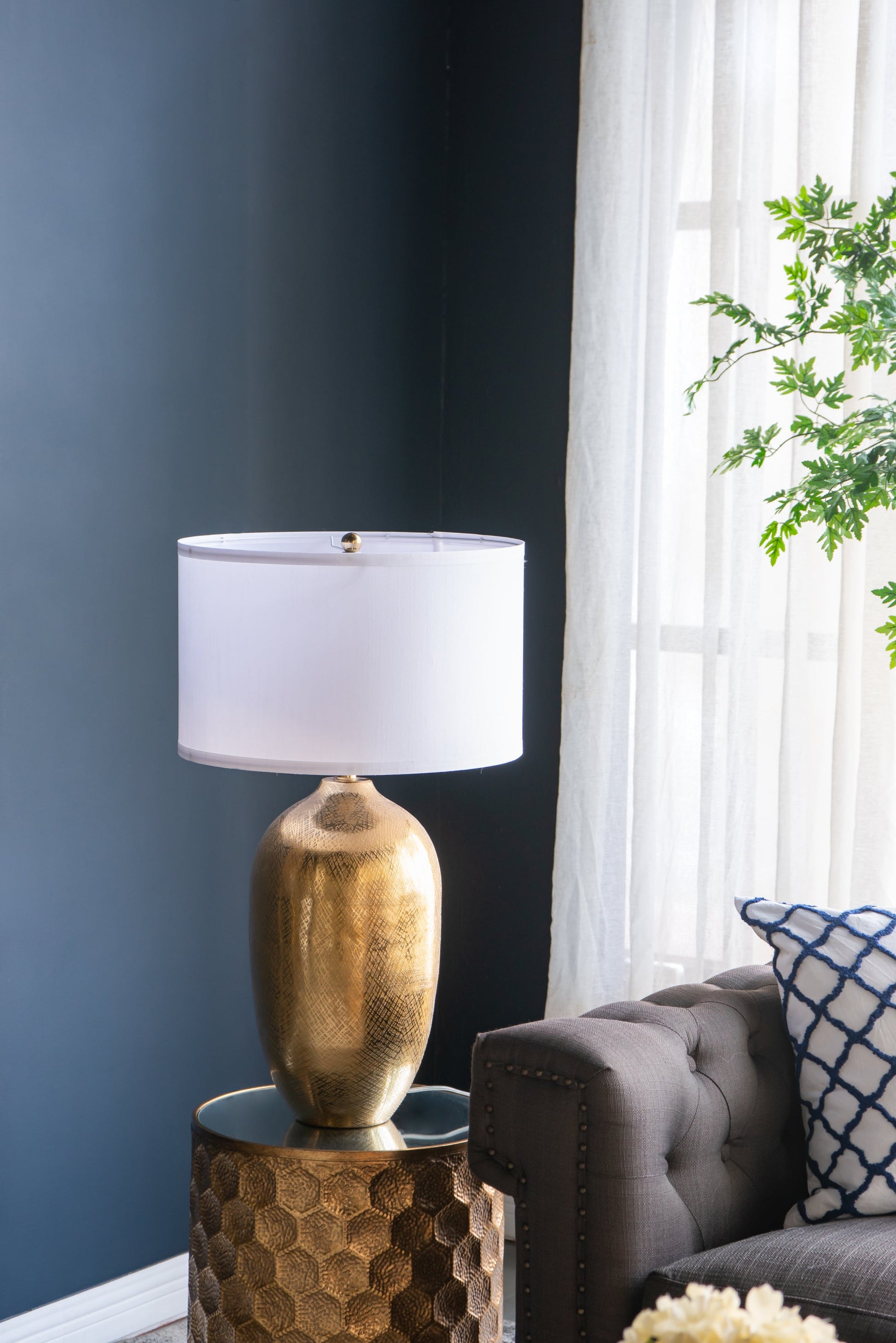 Gold table lamp with white shade on a textured side table next to a gray sofa with a plant in the background.