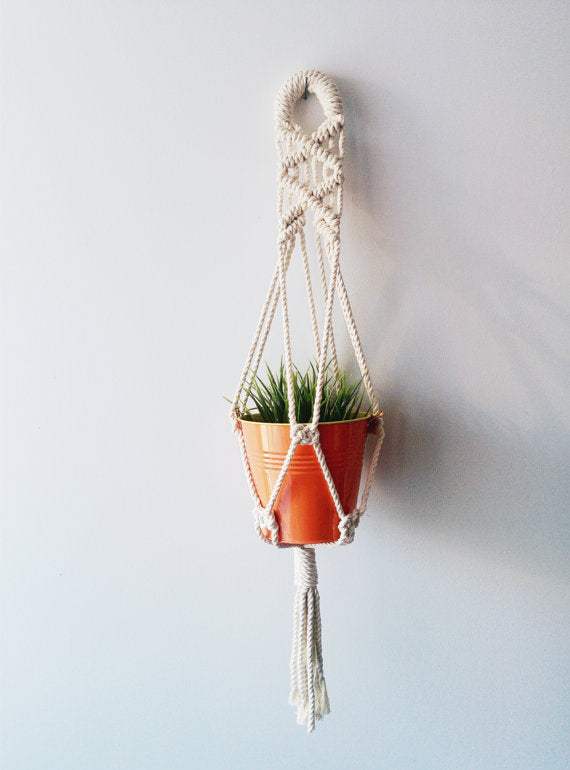 Handmade cotton macrame plant hanger holding an orange pot with a green plant, hanging on a wall.