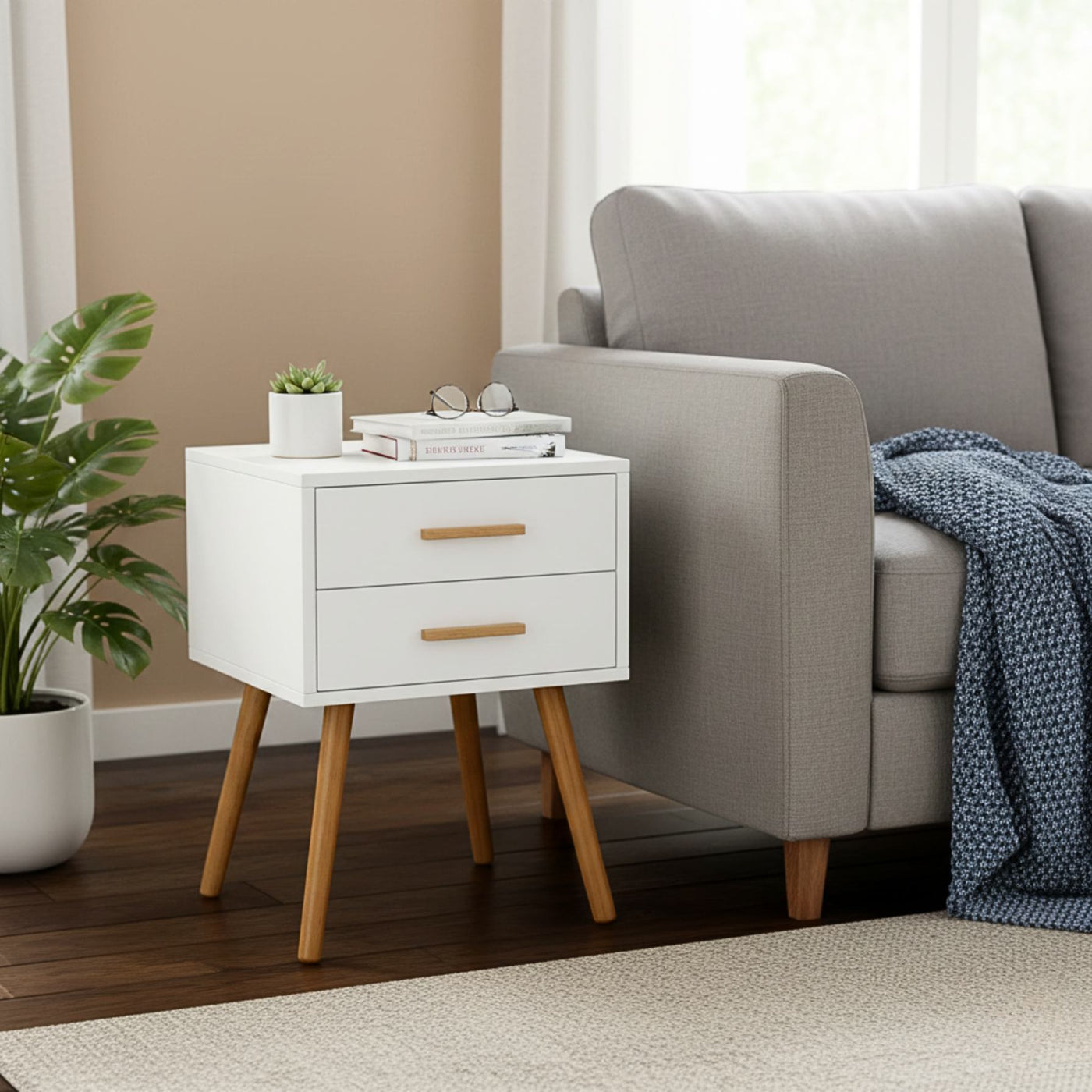 Modern 2-drawer white piano finish end table with wooden legs beside a gray sofa, topped with plant and books.