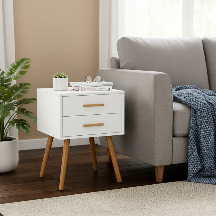 Modern 2-drawer white piano finish end table with wooden legs beside a gray sofa, topped with plant and books.