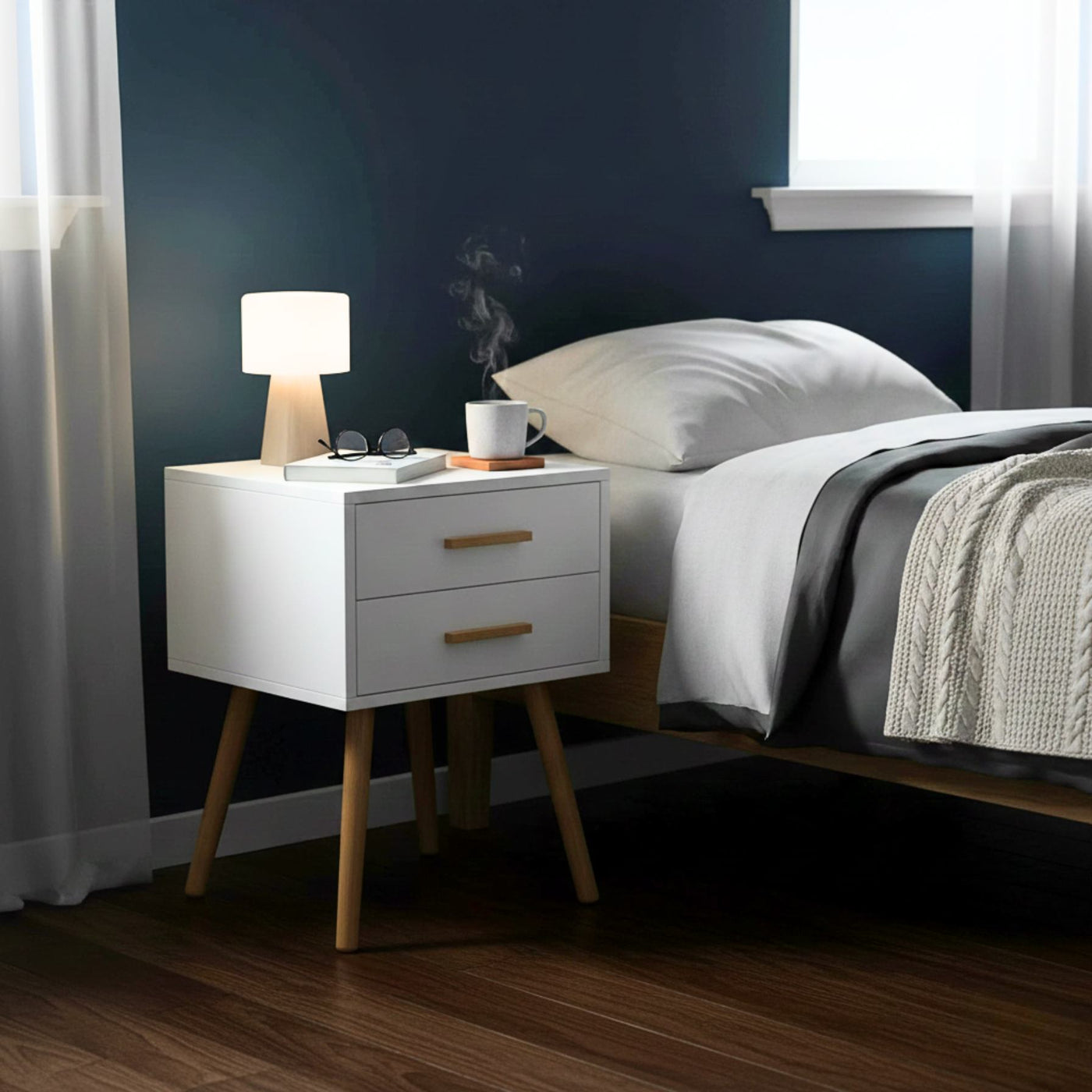 Modern white piano-finish 2-drawer end table with tapered wooden legs beside a bed.