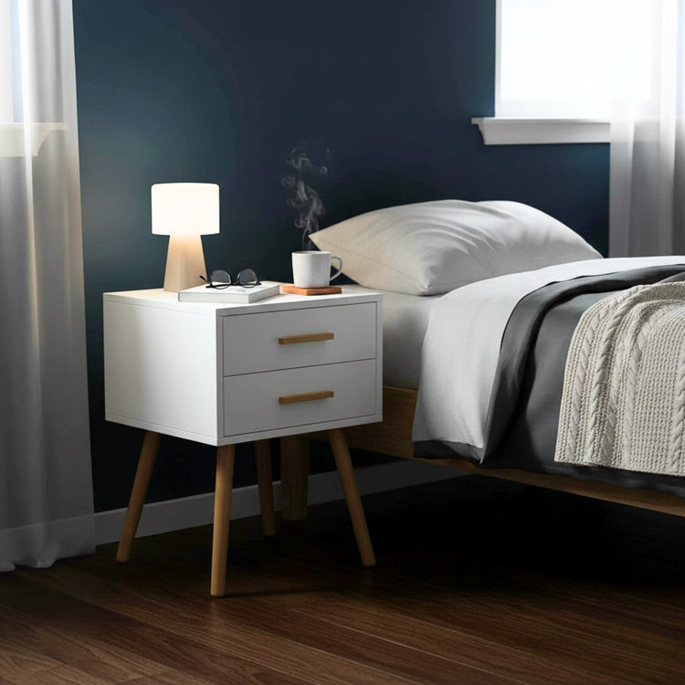 Modern white piano-finish 2-drawer end table with tapered wooden legs beside a bed.