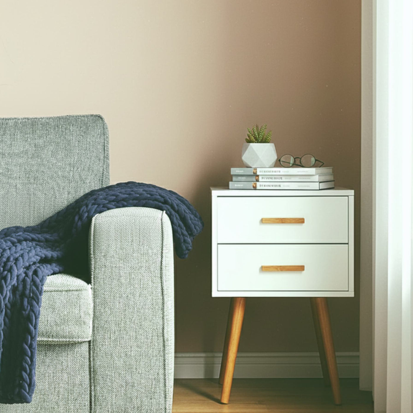 Modern white piano-finish two-drawer end table with wooden legs beside a grey sofa, books and plant.