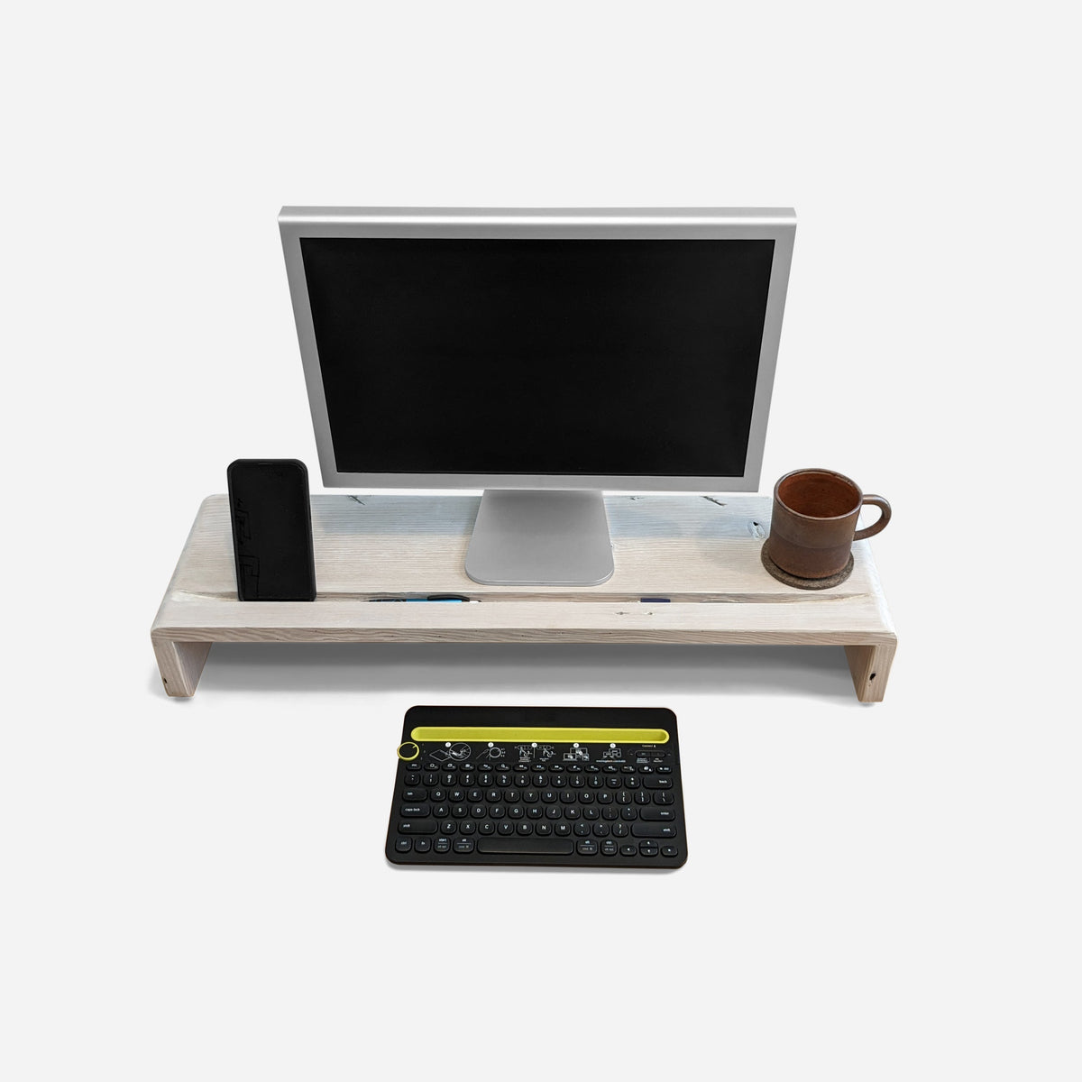 Wood monitor stand in whitewashed finish holding a phone and a stoneware mug, front view on a light studio ground