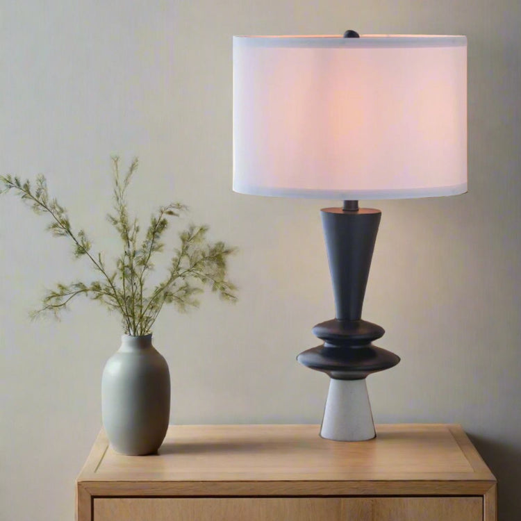 Table lamp with white shade and dark base on a wooden surface next to a vase with greenery.