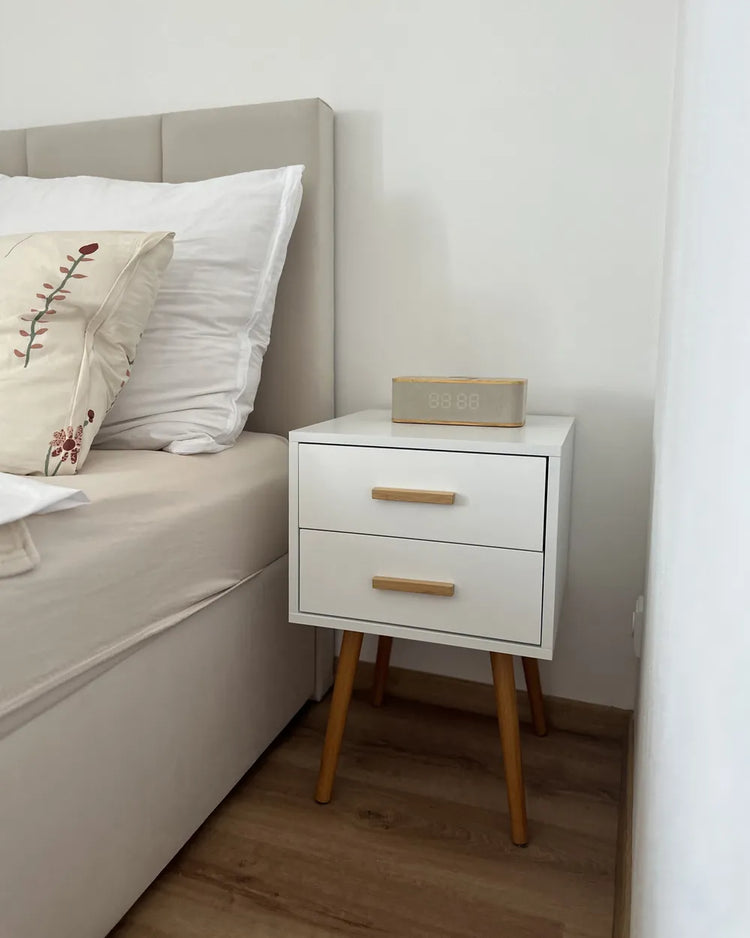 Modern white piano-finish 2-drawer nightstand with tapered wooden legs beside a bed.