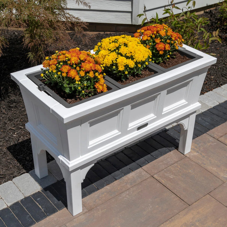 White rectangular raised garden bed planter with removable trays and elevated legs, filled with yellow and orange mums.