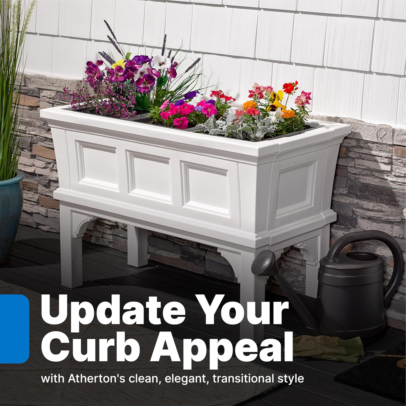 White rectangular raised garden bed planter on legs filled with colorful flowers beside a stone wall.