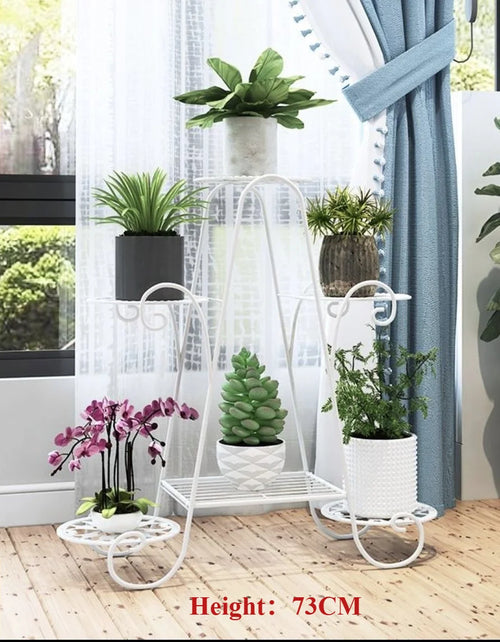 White six-tier metal plant stand on wheels displaying assorted potted plants indoors by a window.
