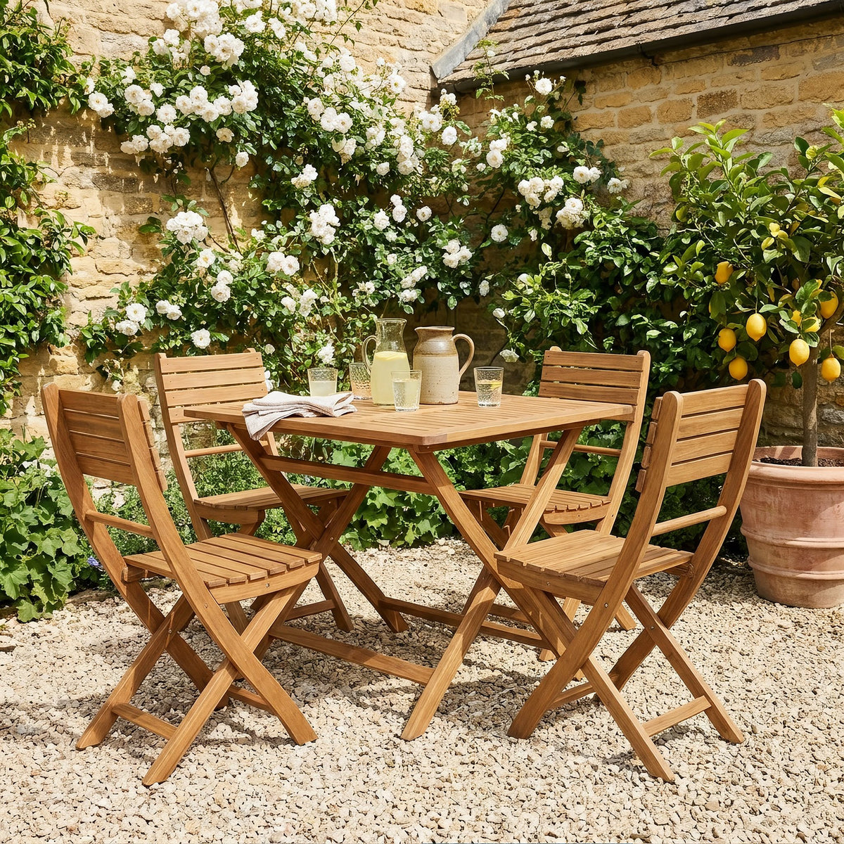 Acacia wood folding dining set with four chairs in a stone courtyard garden with roses and lemonade on the table