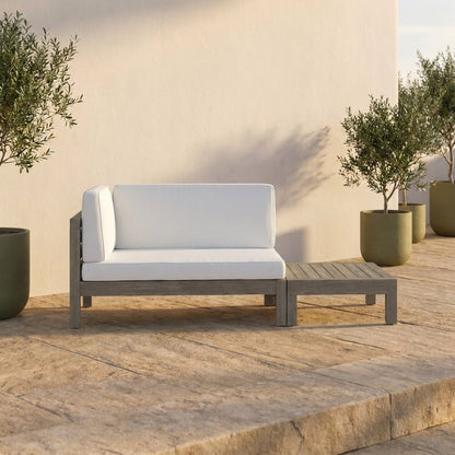 Acacia loveseat and slatted coffee table on a sunlit stone terrace patio beside olive planters at golden hour