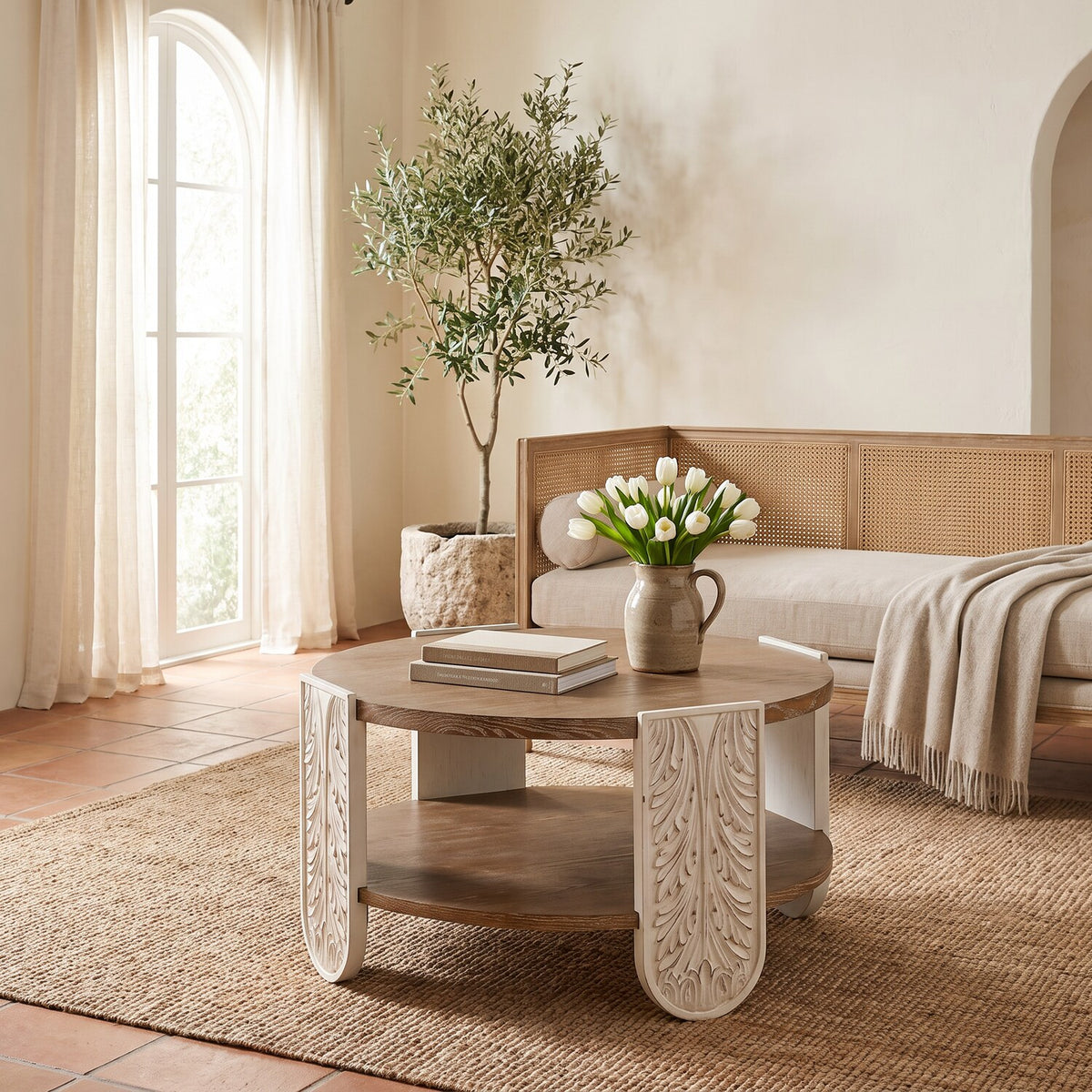Round carved wood coffee table in a sunlit whitewashed sitting room with a daybed and olive tree