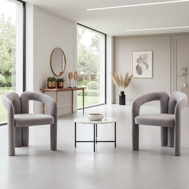 Two gray velvet dining armchairs with curved backs in a bright modern room around a round marble coffee table.