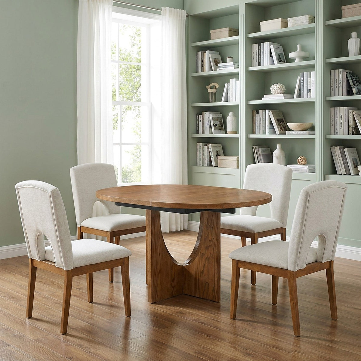 Round wood table with a curved pedestal and four cream upholstered chairs in a pale sage study with built-in bookshelves