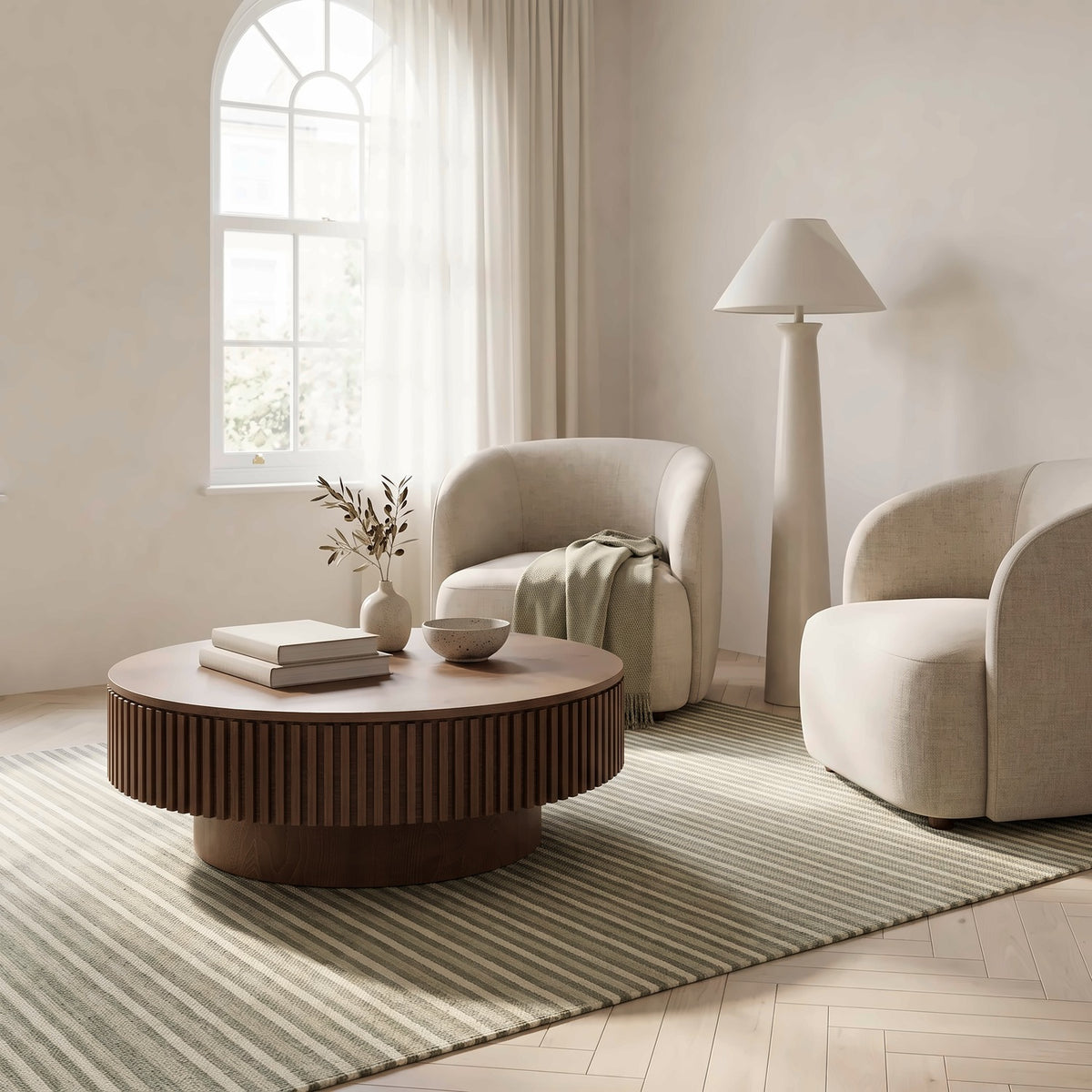 Slatted round walnut table with books, a bowl and a vase beside a tall window, two cream armchairs and a floor lamp behind