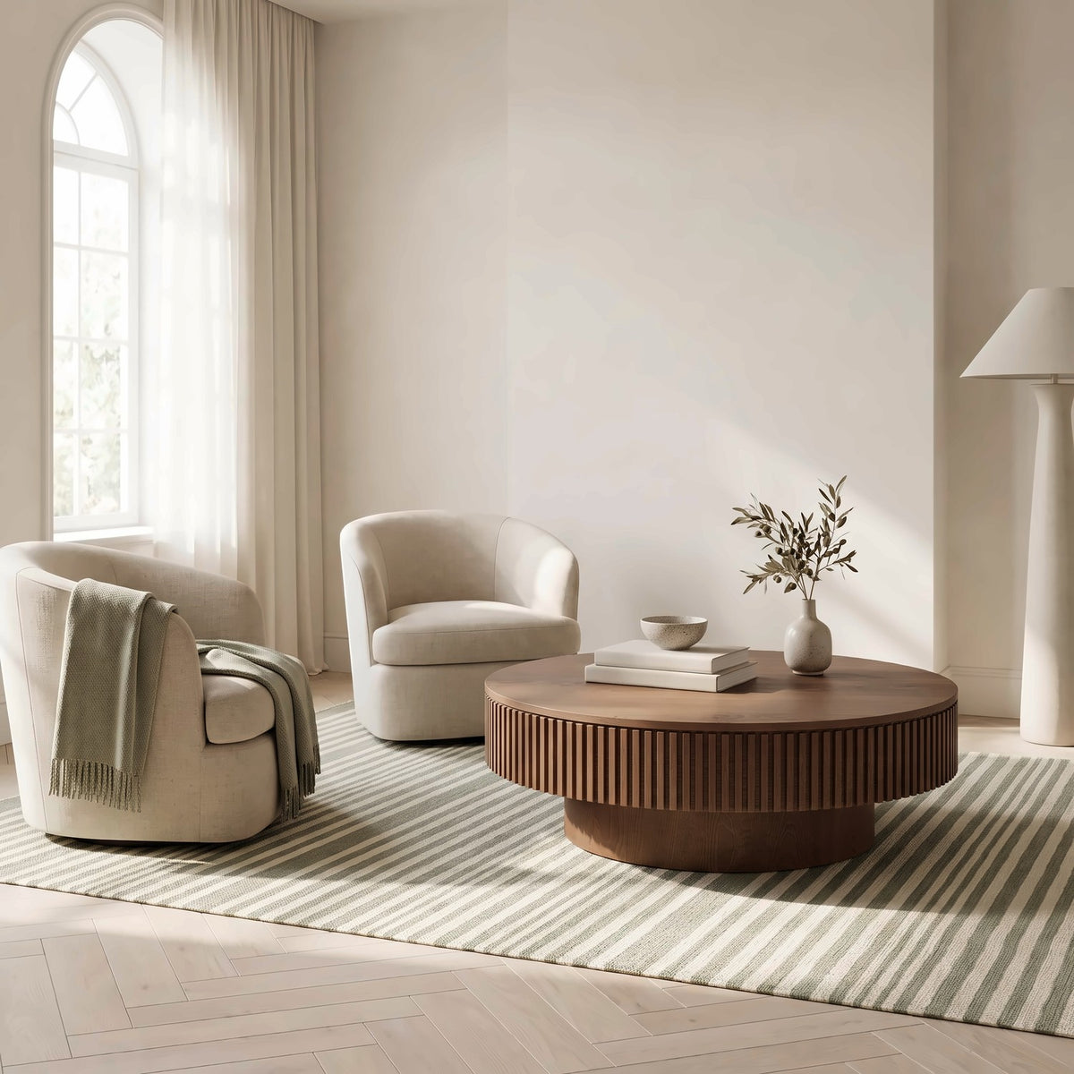 Round walnut table with a slatted drum band between two cream barrel chairs on a striped rug in a bright living room