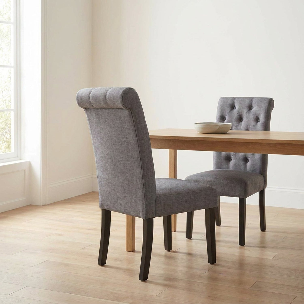 Rolled back of a gray tufted dining chair at an oak dining table by a window