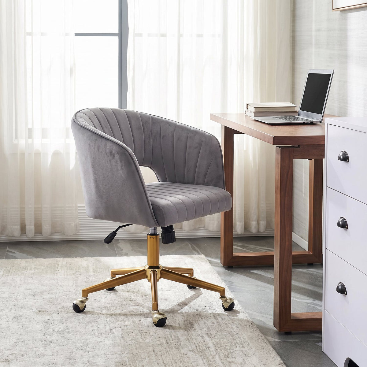 Velvet swivel chair beside a walnut desk with an open laptop by sheer-curtained windows in a home office