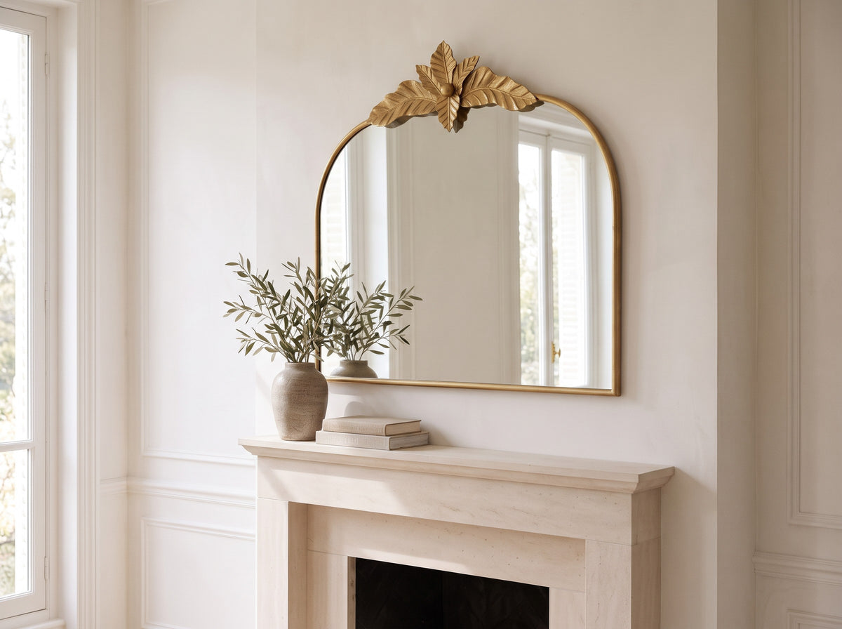 Gold arched mirror seen at an angle over a mantel with books and a stone vase of olive branches