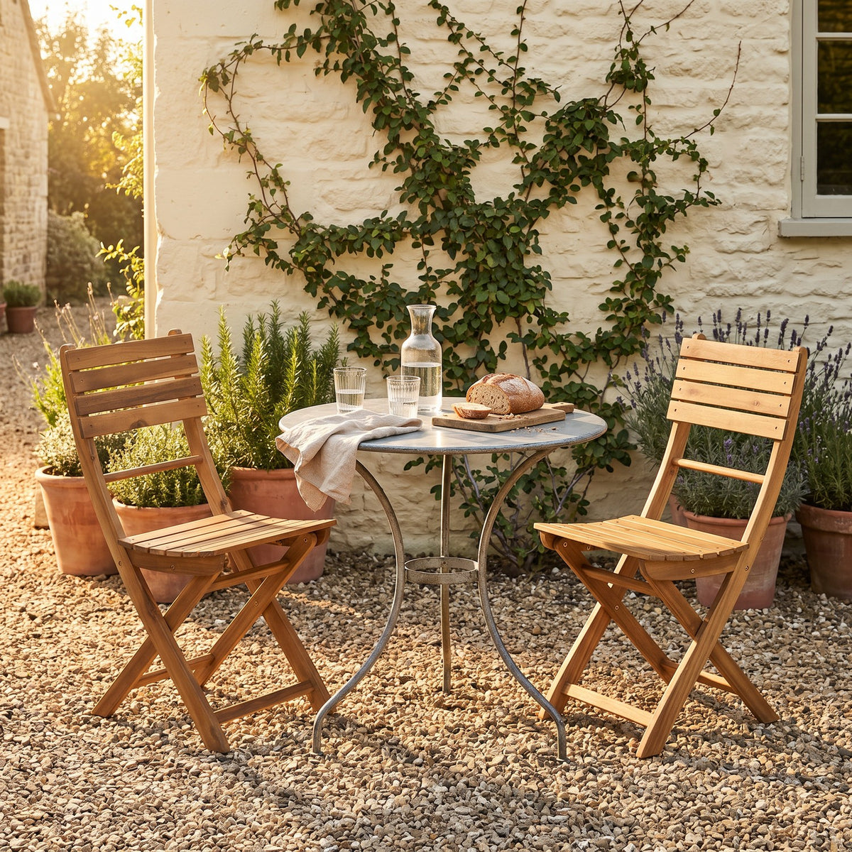 Two acacia folding chairs at a zinc bistro table in a gravel cottage courtyard, rosemary and lavender pots behind