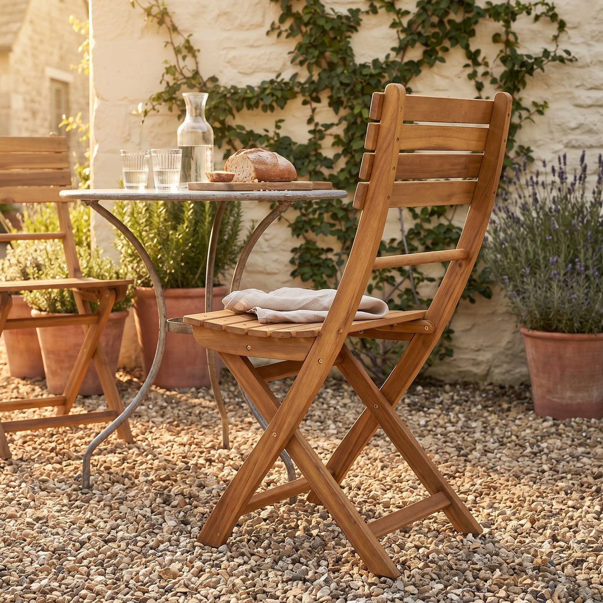 An acacia folding chair in a cottage courtyard at golden hour, linen napkin on the seat and the bistro table behind