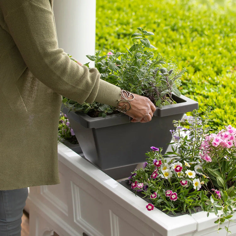 Hands placing herb plants into a rectangular raised garden bed planter on a porch.