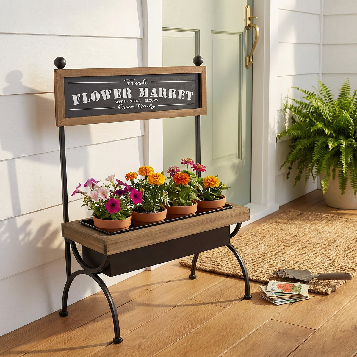 Flower market stand with potted petunias and marigolds on a sunny front porch beside a sage green door and a coir doormat