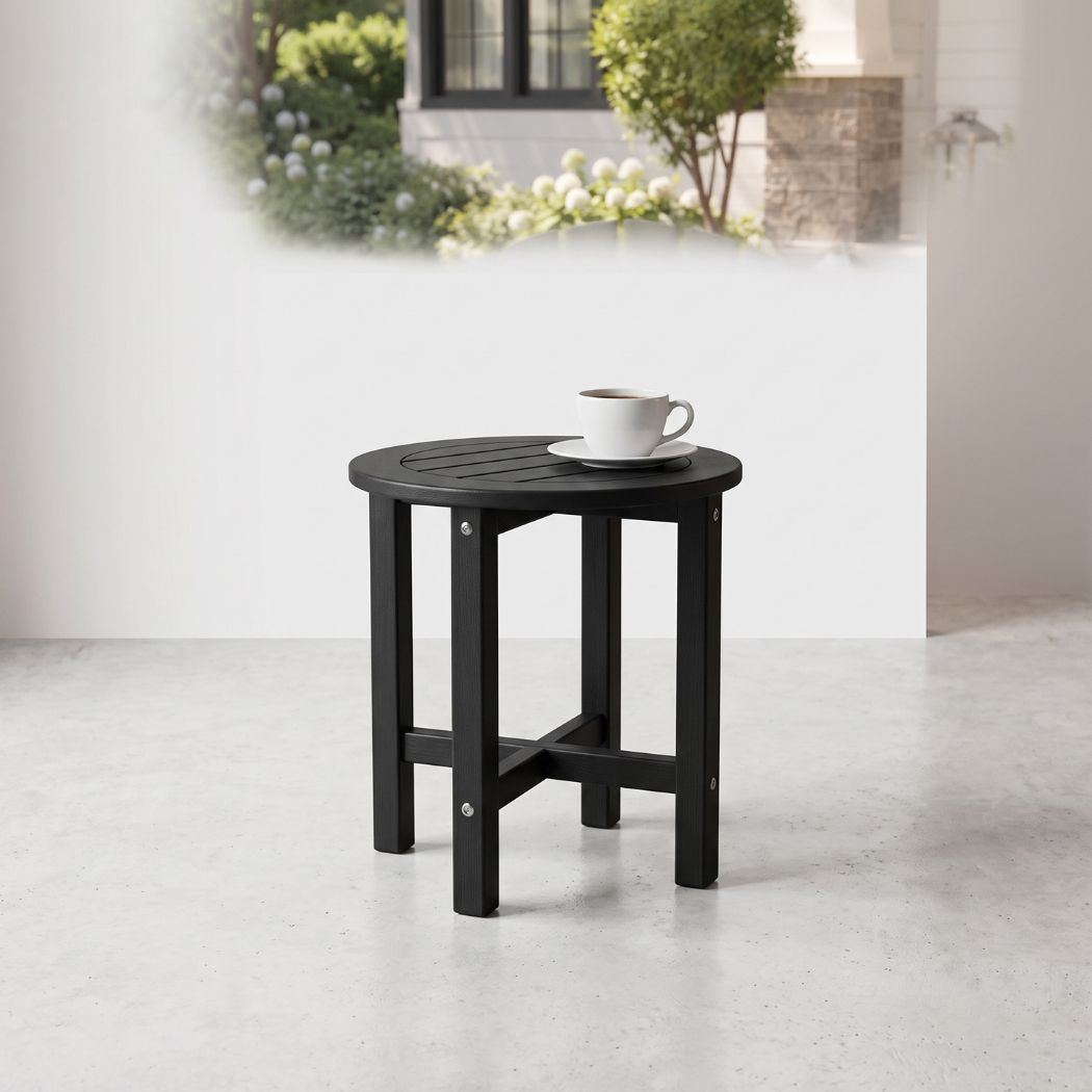 A small round black outdoor side table with a slatted top and four legs, shown on a light concrete floor against a white wall. A white cup and saucer sit on the table.