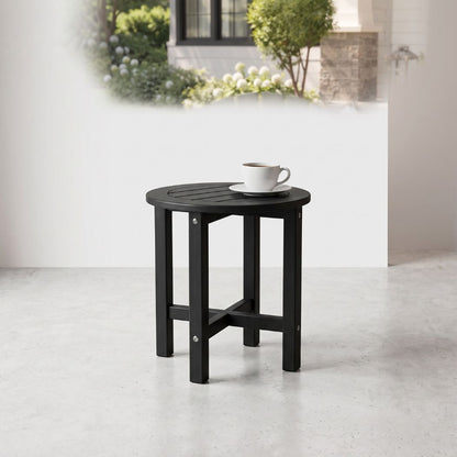 A small round black outdoor side table with a slatted top and four legs, shown on a light concrete floor against a white wall. A white cup and saucer sit on the table.
