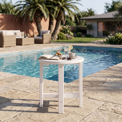 A small round white outdoor side table with legs stands on a stone patio beside a swimming pool. An open book, a cup of coffee, a drink with a lemon slice, and a small candle are arranged on the tabletop.