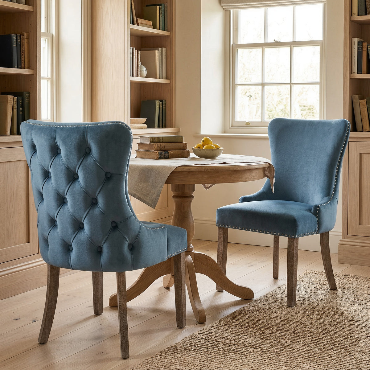 Close view of light blue tufted velvet wingback dining chairs at a round oak table by a bright window