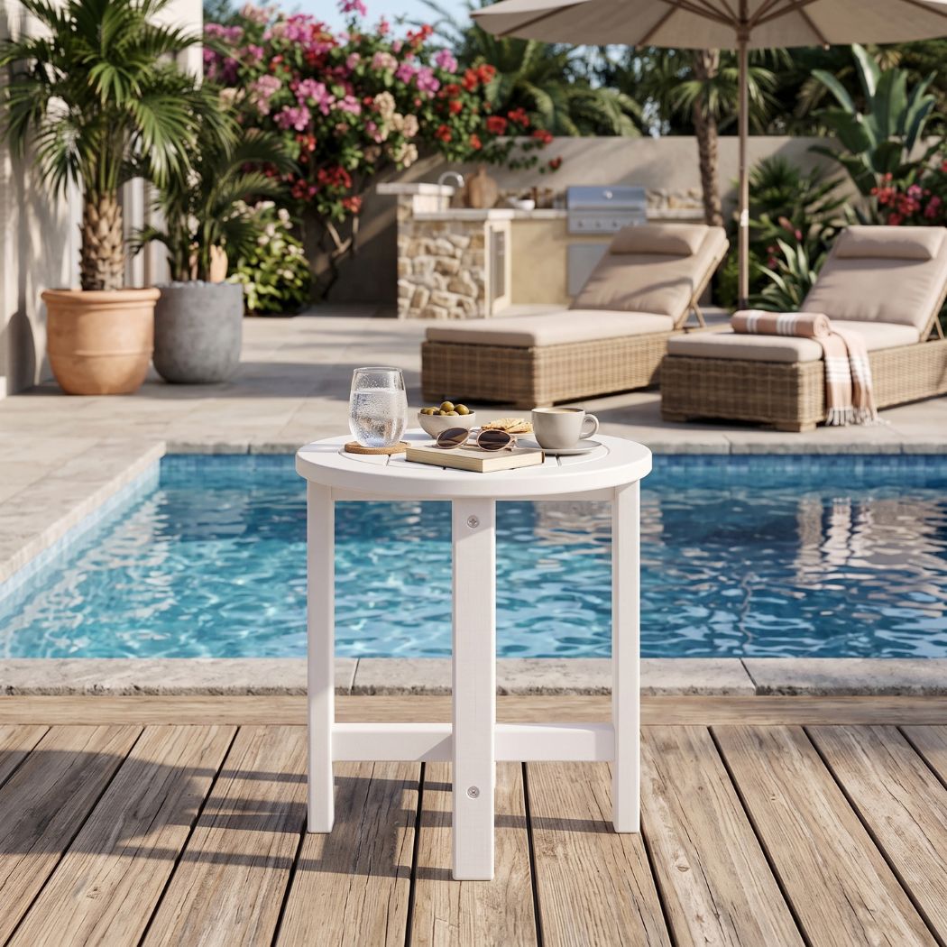 A small round white outdoor bar table with legs, shown on a wooden deck beside a swimming pool. The table is styled with a glass, a book, and a small cup on top.
